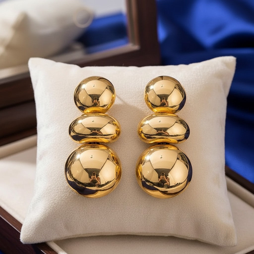 House of eLLe I'm Flexible gold drop earrings featuring three stacked polished spheres on a white display cushion.