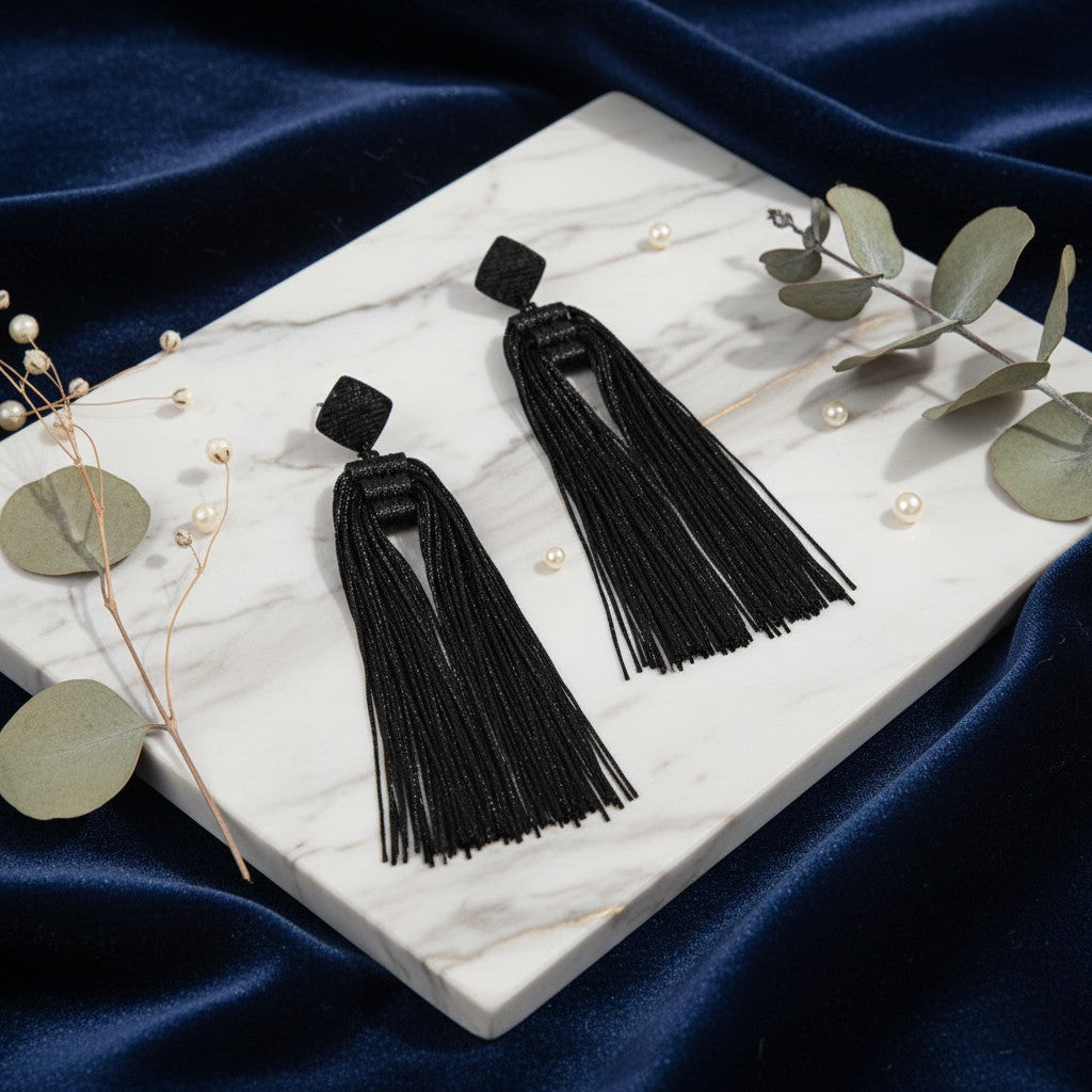 House of eLLe Queen Era black tassel earrings with diamond-shaped studs on a marble display with eucalyptus leaves.