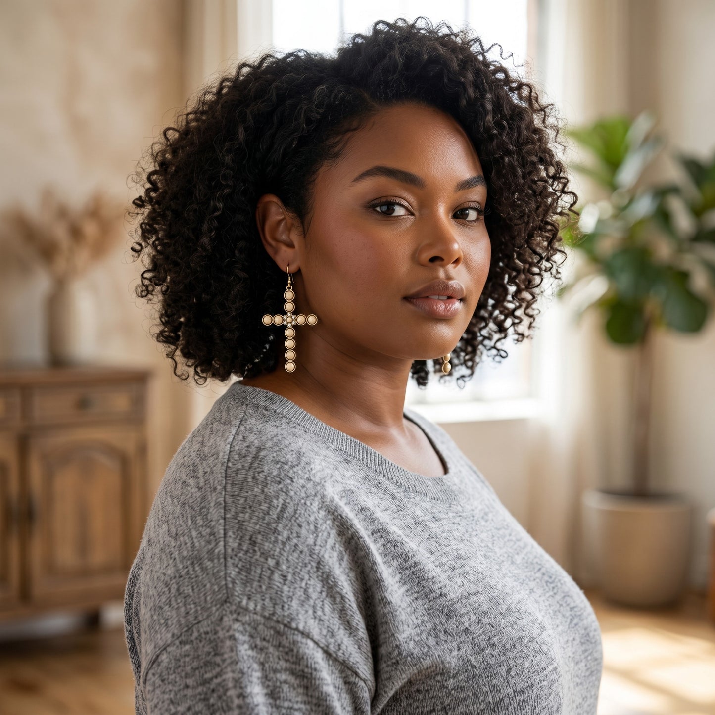 Woman wearing House of eLLe cream bling cross earrings with a grey sweater in a bright, cozy living room.