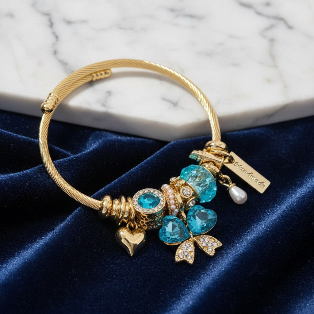 House of eLLe gold stainless steel charm bracelet featuring blue crystal hearts, a bow, and a pearl charm.