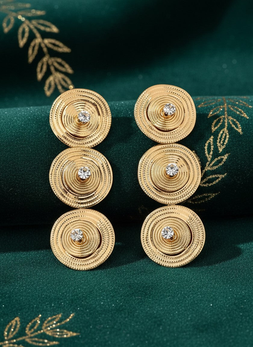 House of eLLe gold-tone triple circular drop earrings with central crystal accents on a dark green fabric background.
