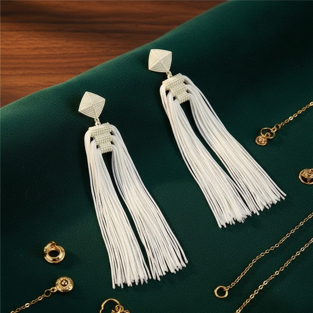 House of eLLe Queen Era white beaded tassel earrings with pyramid stud tops on a dark green fabric background.